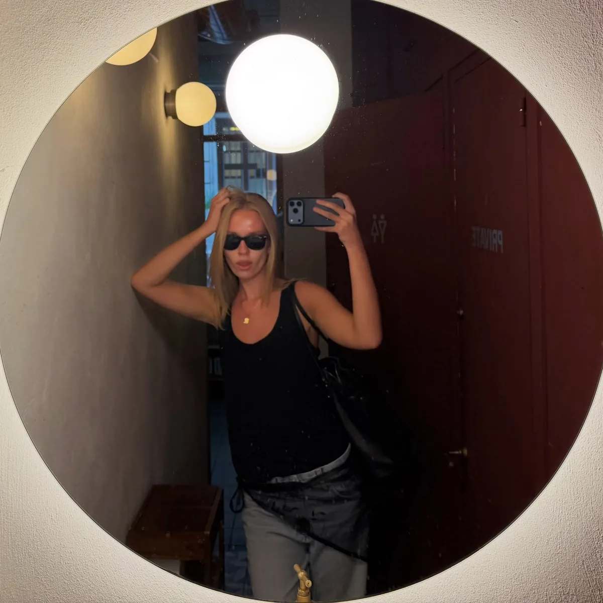 sofia in sunglasses and a black tank top, taking a photo in a round lit mirror