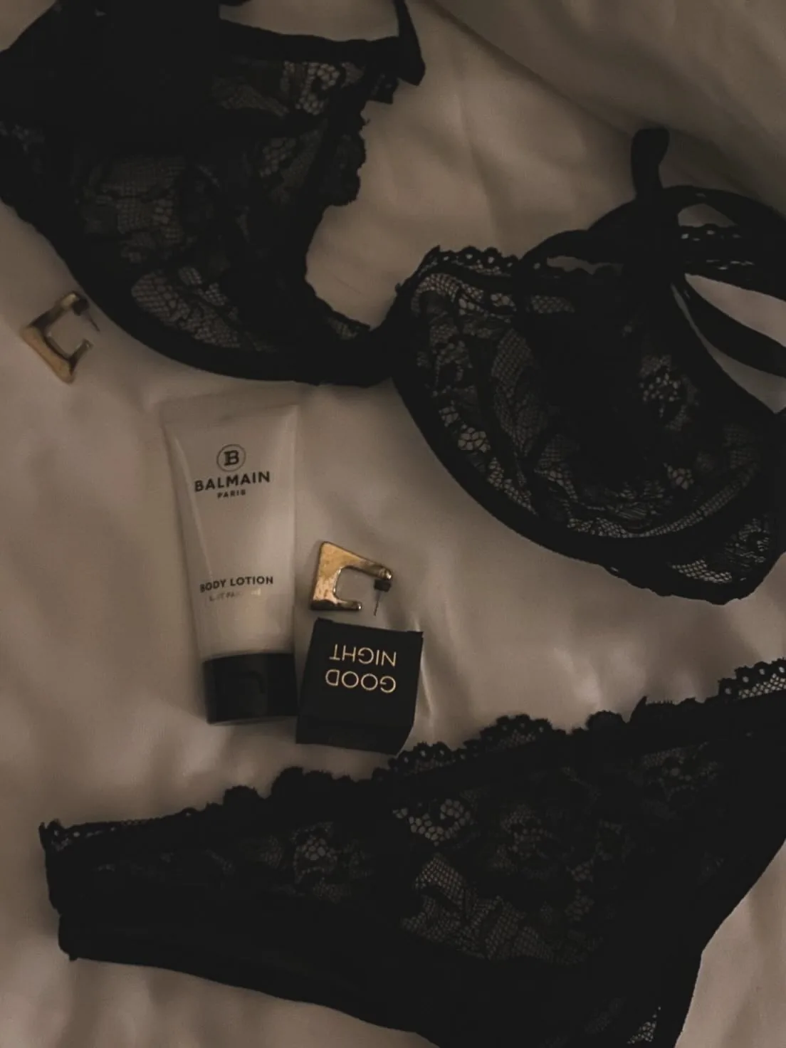 Black lace lingerie, Balmain body lotion and a Good Night candle on white sheets