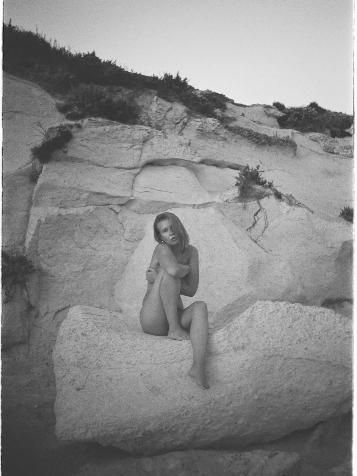 sofia sitting on white rocks by the sea, arms around her knees, black and white film photo