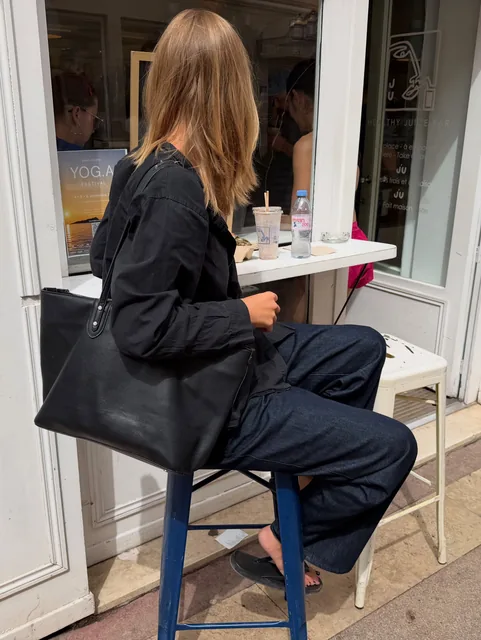 sofia on a stool at a café window, black shirt and jeans, a large black tote